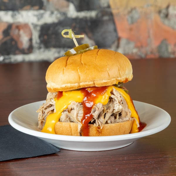 Pulled pork sandwich with melted cheese, barbecue sauce, and pickles on a white plate.