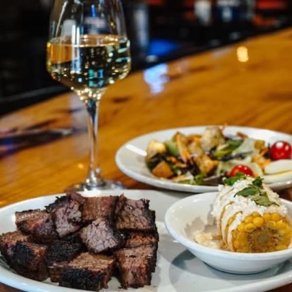 Plates of brisket, street corn, and salad with a glass of white wine on a wooden bar.