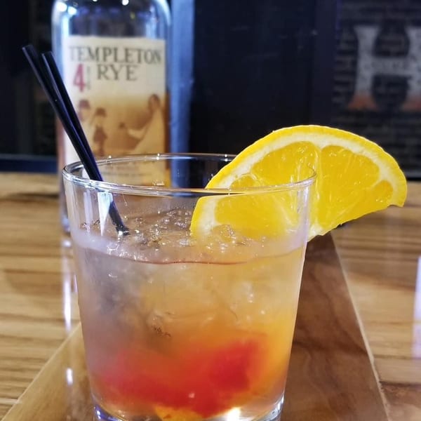 An Old Fashioned cocktail with an orange slice and two black straws, with a bottle of Templeton Rye in the background.