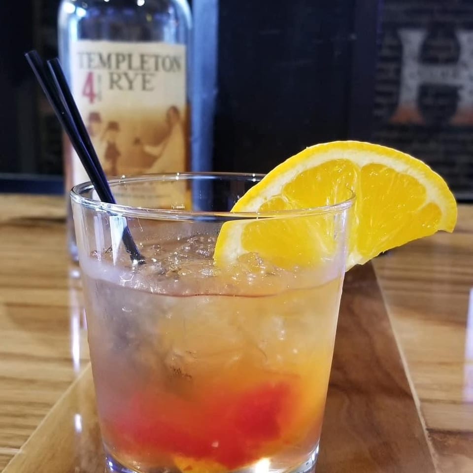 An Old Fashioned cocktail with an orange slice and two black straws, with a bottle of Templeton Rye in the background.