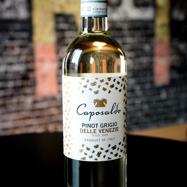 A bottle of Caposaldo Pinot Grigio Delle Venezie white wine, with a gold label, against a brick wall.