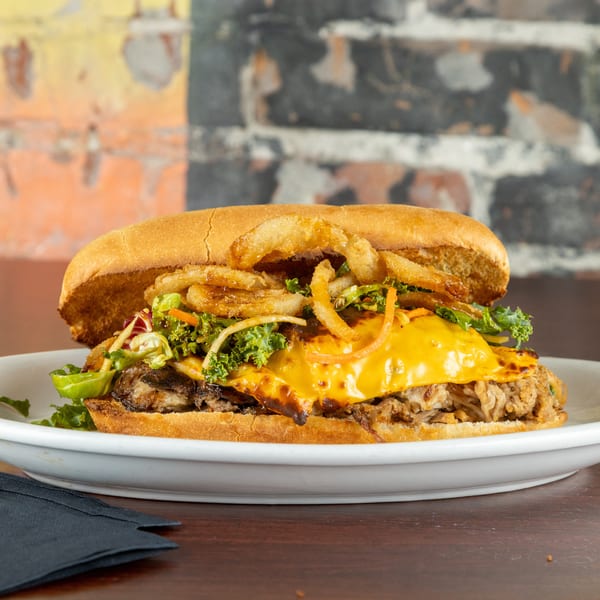 A pulled pork sandwich with melted cheese, kale, and crispy fried onions on a white plate.
