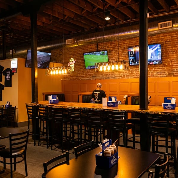 Spacious restaurant interior with a long wooden bar, tables, brick walls, and TVs showing sports.