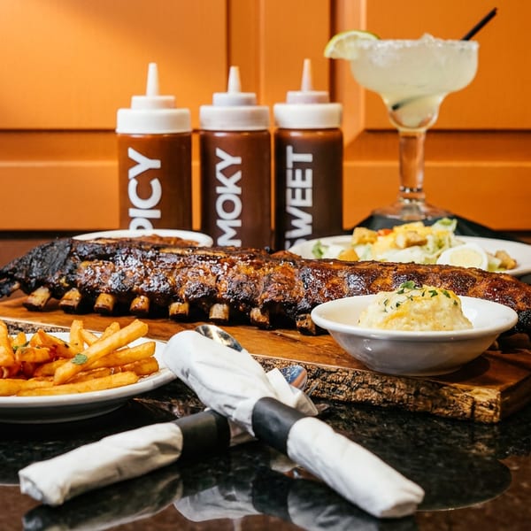 Rack of ribs, fries, mashed potatoes, salad, three sauces, and a margarita on a dark table.