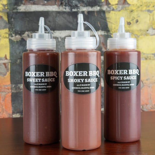 Three bottles of Boxer BBQ sauce: Sweet, Smoky, and Spicy, on a wooden table.