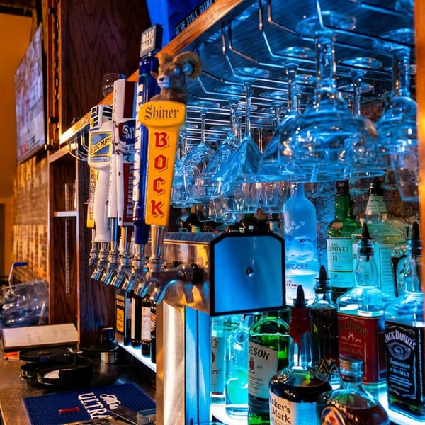 Bartender working behind a bar with beer taps, liquor bottles, and hanging glasses under blue light.