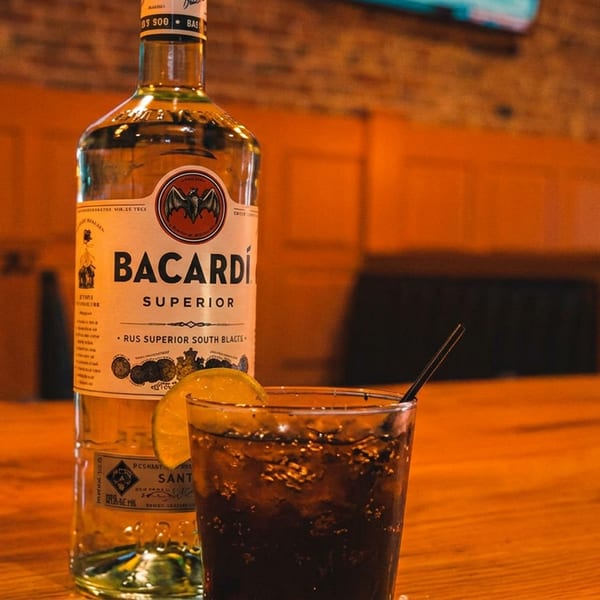 Bacardi Superior bottle and a rum and coke with lime on a wooden bar table.