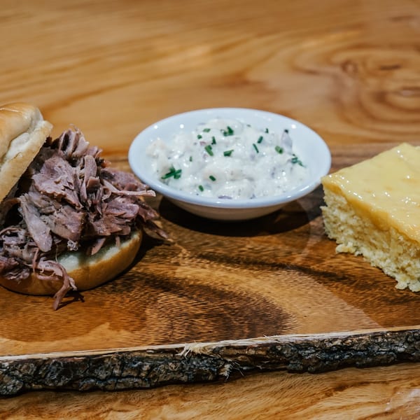 Pulled pork sandwich, potato salad, and cornbread on a rustic wooden board.