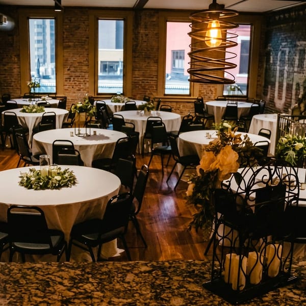 Spacious event room with round tables, buffet, exposed brick walls, and industrial pendant lights.