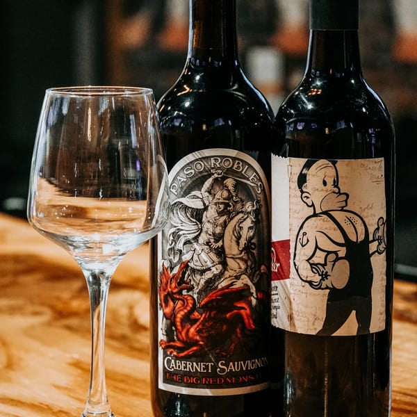 Two wine bottles, one with a knight and dragon label, the other with a cartoon character, next to a wine glass.