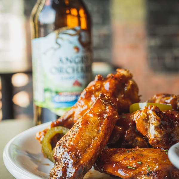 A plate of glistening BBQ chicken wings with jalapeños and a bottle of Angry Orchard cider in the background.