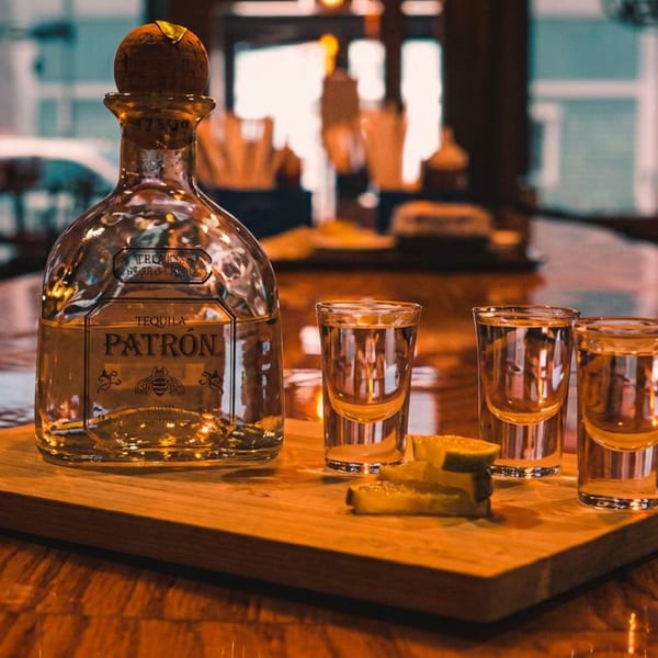 Patron tequila bottle, three full shot glasses, and lime wedges on a wooden board at a bar.
