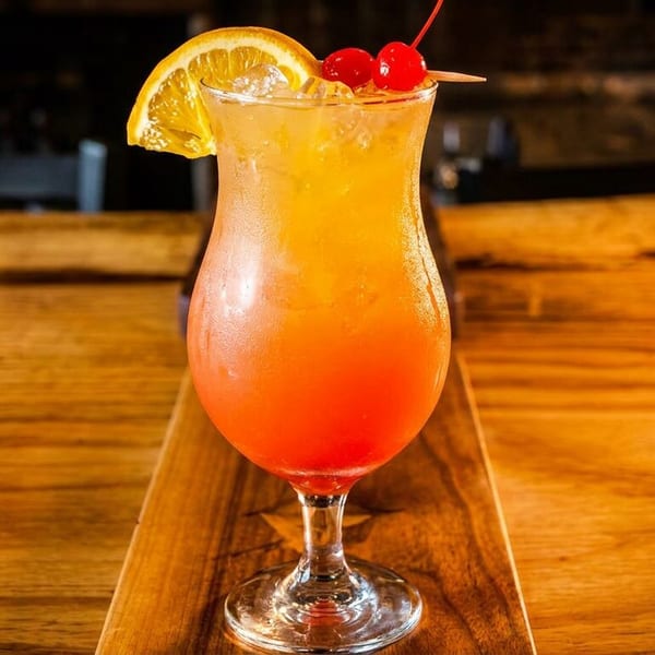Orange and red cocktail in a hurricane glass with orange slice and cherries on a wooden table.