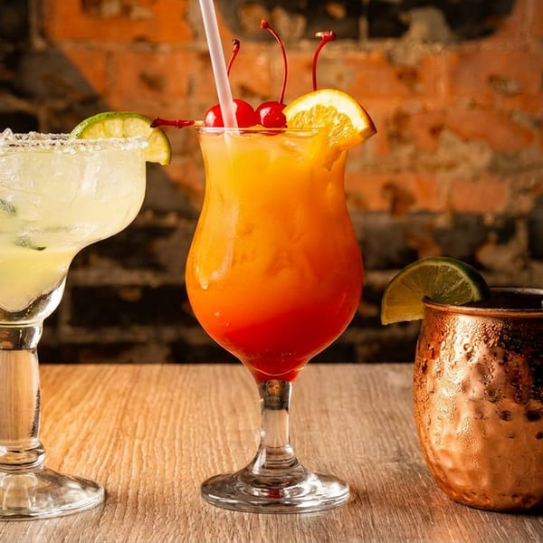 Three vibrant cocktails: a margarita, a layered orange drink, and a Moscow Mule on a wooden table.